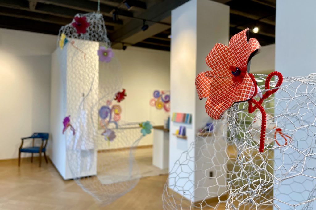 A photo of a gallery installation with sculptures made of white wire mesh and colorful fabric flowers. In the background, there are abstract art pieces and shelves with other art displays.