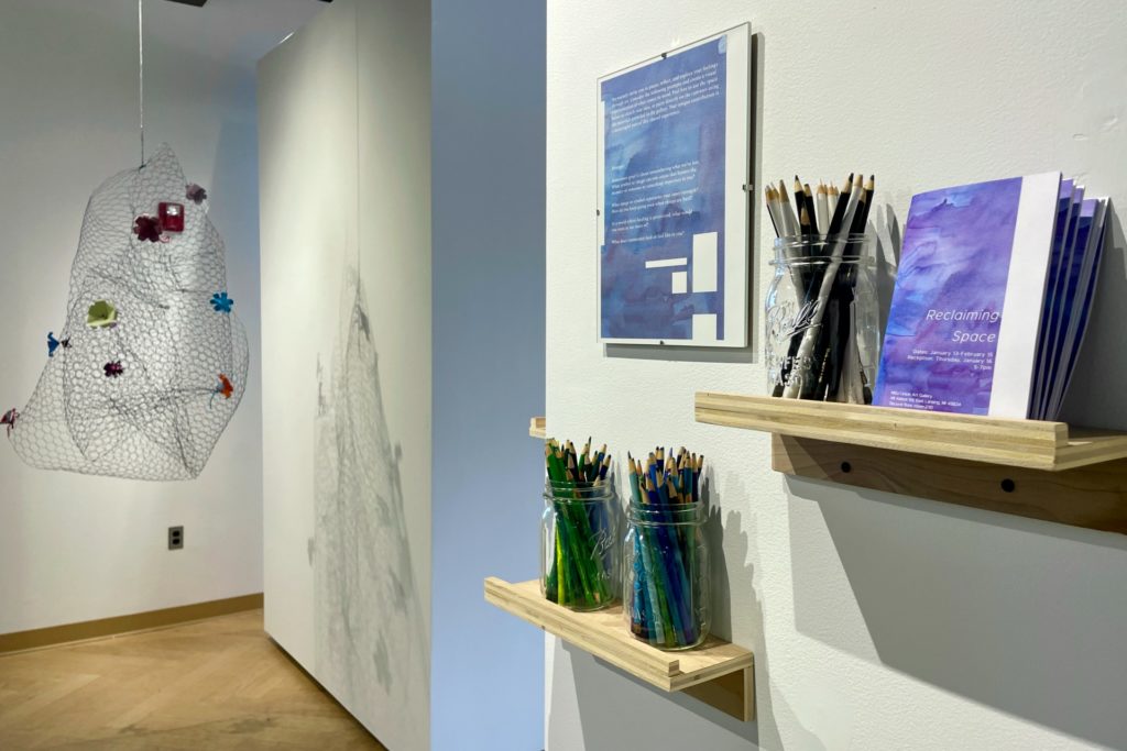 A picture of a gallery exhibit, with shelves mounted on a gallery wall holding glass jars filled with colored pencils and black markers. A flyer for the "Reclaiming Space" exhibit and a framed description are displayed on the wall. A wire mesh sculpture with fabric flowers hangs in the background.