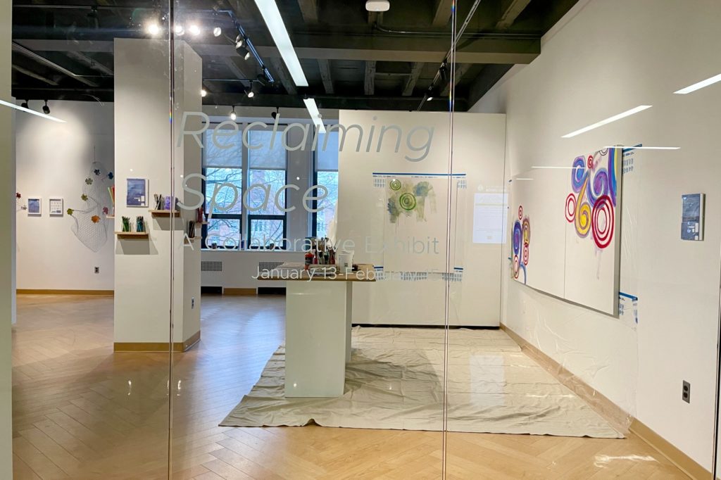 A wide view photography of the "Reclaiming Space" gallery exhibit through a glass door. The space includes abstract circular paintings, wire mesh sculptures with floral details, and art materials displayed on shelves. Text on the glass reads, "Reclaiming Space: A Collaborative Exhibit, January 13 - February 15."