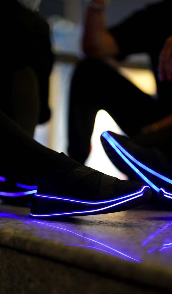 A close-up of black sneakers with glowing blue LED lighting along the soles in a dim environment. 