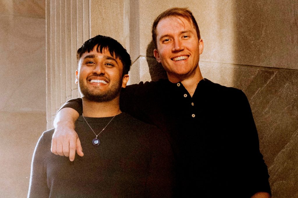 A picture of two individuals standing close together, smiling warmly. One person has an arm around the other's shoulder. They are dressed casually, with one wearing a necklace featuring an eye-shaped pendant. A stone wall and columns provide the backdrop.