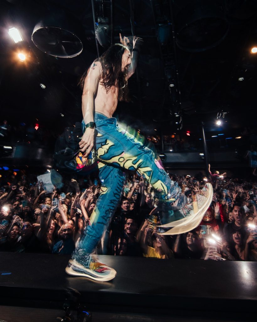 A photo of a performer with long hair and colorful jeans strides across a stage during a live concert. The crowd, holding phones and cheering, is visible in the background. The performer wears vibrant sneakers with bold patterns a white LEDs. 