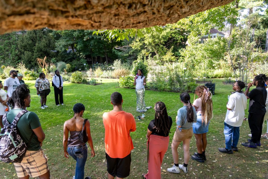 A group of people standing in a semicircle on a grassy area inside Beal Garden with a woman who is standing at the end of the semicircle holding a microphone and speaking. The rest of the group is looking at her. 