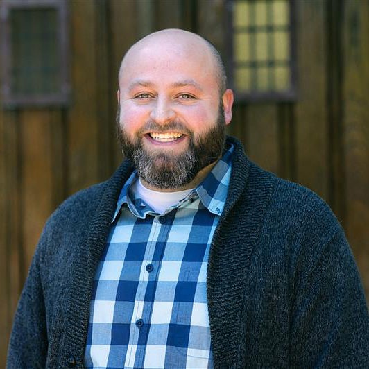  A man with a beard and shaved head smiles. He is wearing a blue plaid shirt and a gray cardigan, standing in front of a wooden building with small windowpanes.