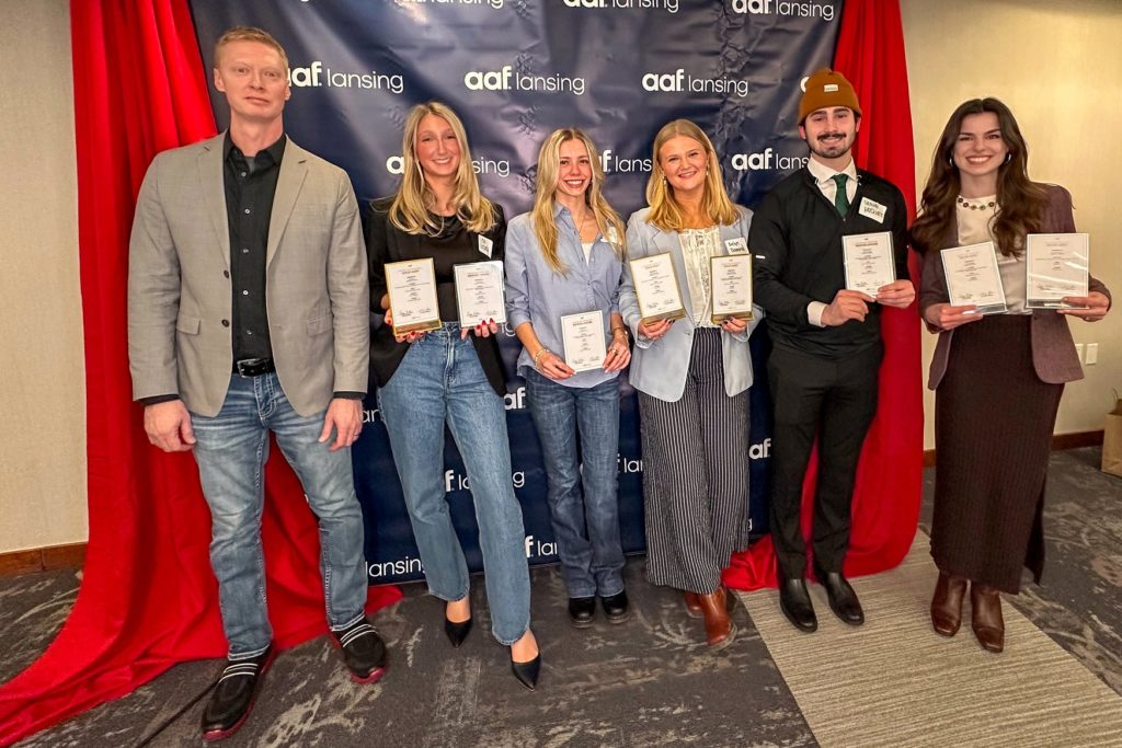 Six individuals stand together in front of a navy blue backdrop with "AAF Lansing" logos, holding award plaques. A red curtain frames the backdrop. They are dressed in business and semi-formal attire, smiling at the camera.