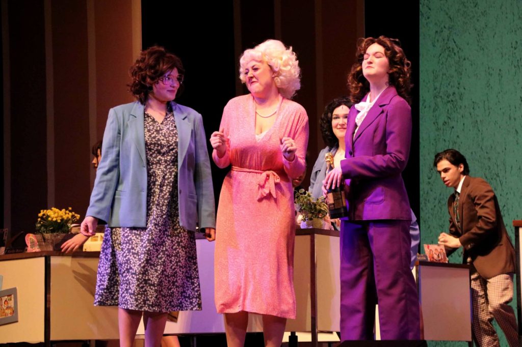 Three woman on stage wearing brightly covered office wear on the set of an office. 