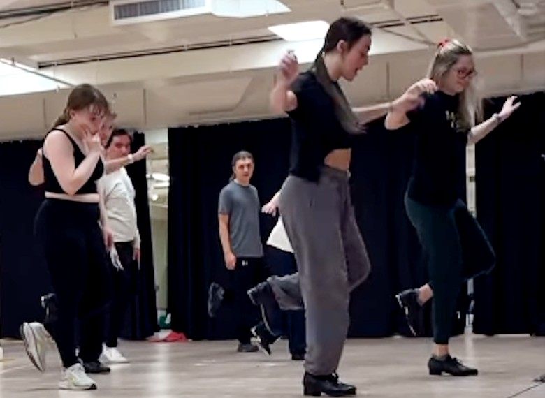 A group of performers rehearses a tap dance routine in a studio with black curtains in the background. 