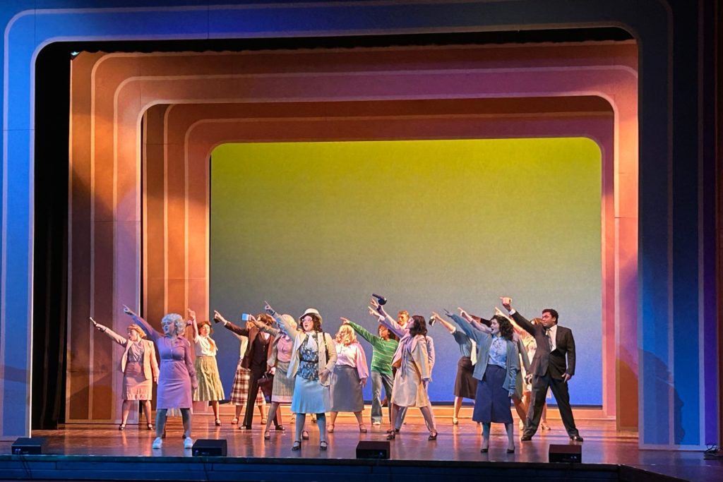 A staged dance number with a number of people whoare dressed for the office. All of them are standing and pointing one hand in the air. 