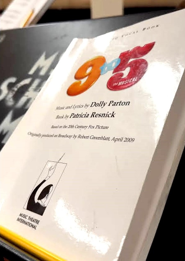 A close-up of a script book for 9 to 5: The Musical, with the title in bold, colorful letters on the cover. Below the title, text credits Dolly Parton for music and lyrics and Patricia Resnick for the book. The script rests on a black music stand, with a blurred background.