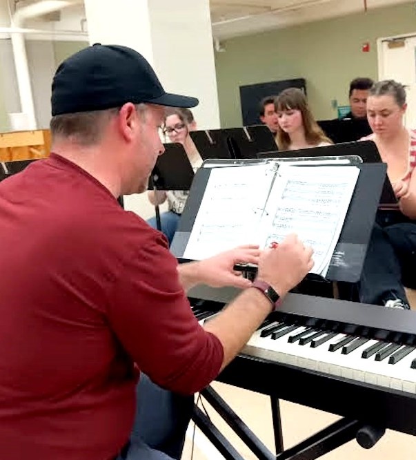 A man wearing a black cap and red shirt sits at a keyboard, marking notes in sheet music on a stand. In the background, a group of students, some holding scripts, sit in chairs.