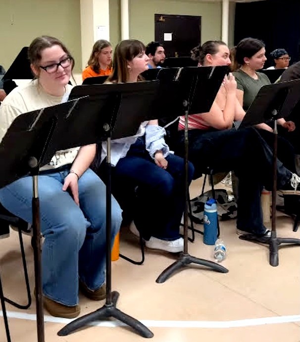 A group of students sits in a rehearsal room. They are seated behind black music stands, with some resting their hands on their laps or holding scripts. 