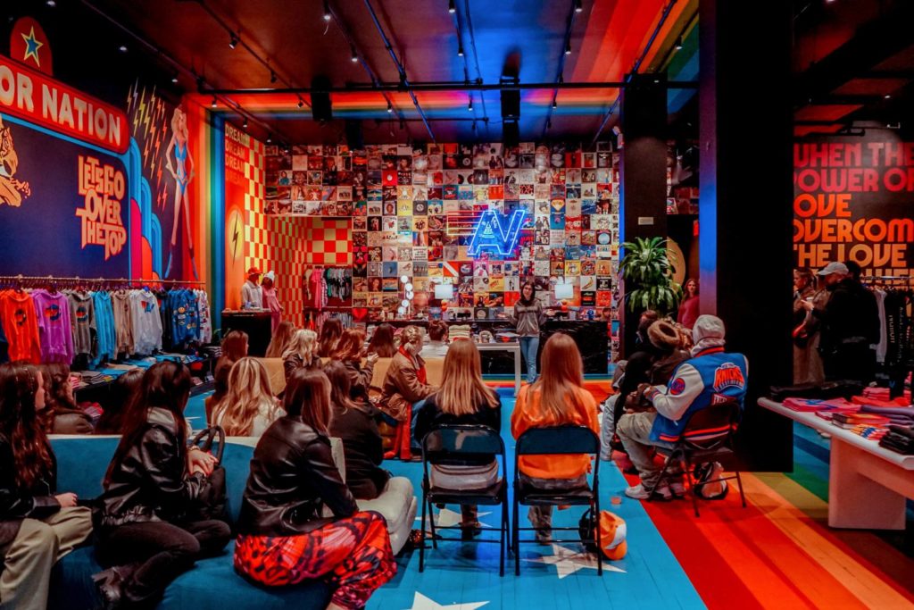 A group of people sits in folding chairs inside a vibrant, colorful retail space with walls covered in neon signs, posters, and clothing displays. A speaker stands at the front with a glowing blue "AV" logo behind them