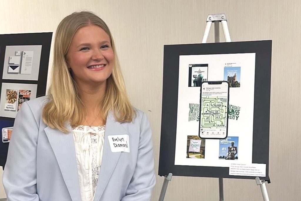 A young woman with blonde hair, wearing a light blue blazer and a name tag reading "Evelyn Diamond," smiles while standing next to a display board featuring a mobile phone design and various promotional graphics.