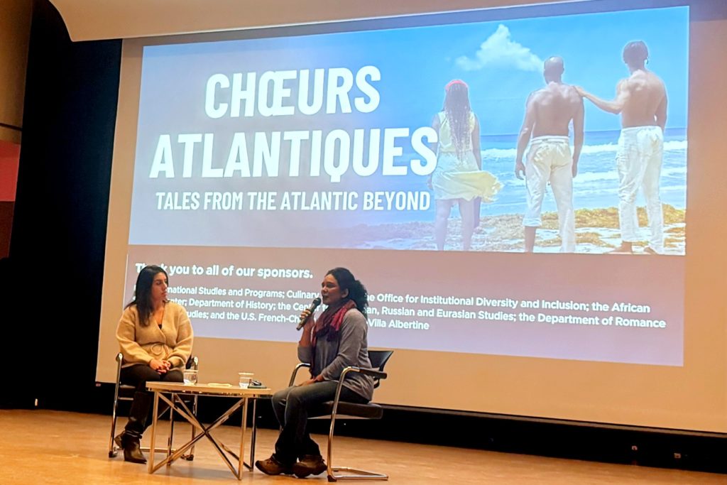 Two women sitting on a stage. One of the women is holding a microphone and speaking into it. Behind them is a large screen that is has an image projected onto it with the words  the words: "Chœurs Atlantiques Tales from thr Atlantic Beyond."