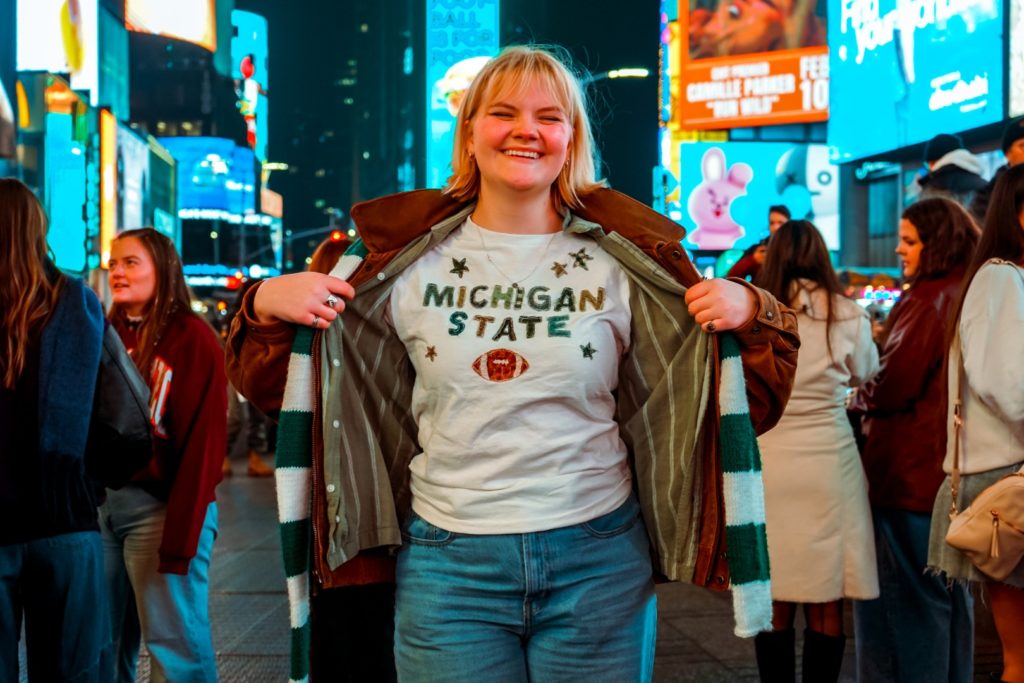 A young woman with blonde hair smiles while standing in Times Square at night. Bright billboards and a crowd of people surround her.