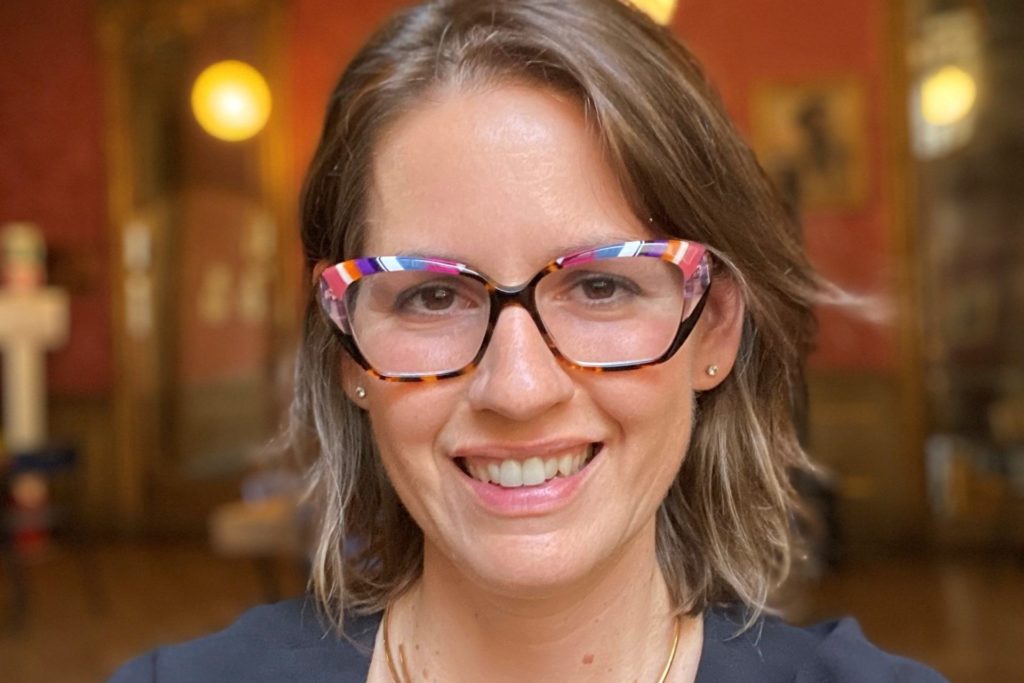 A women wearing colorful glasses and a gold necklace smiles.