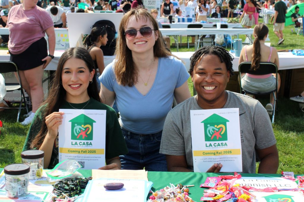 Three students sit at a table at an outdoor campus event, holding flyers for "La Casa." The table is covered with informational materials and candy, and other event attendees are visible in the background