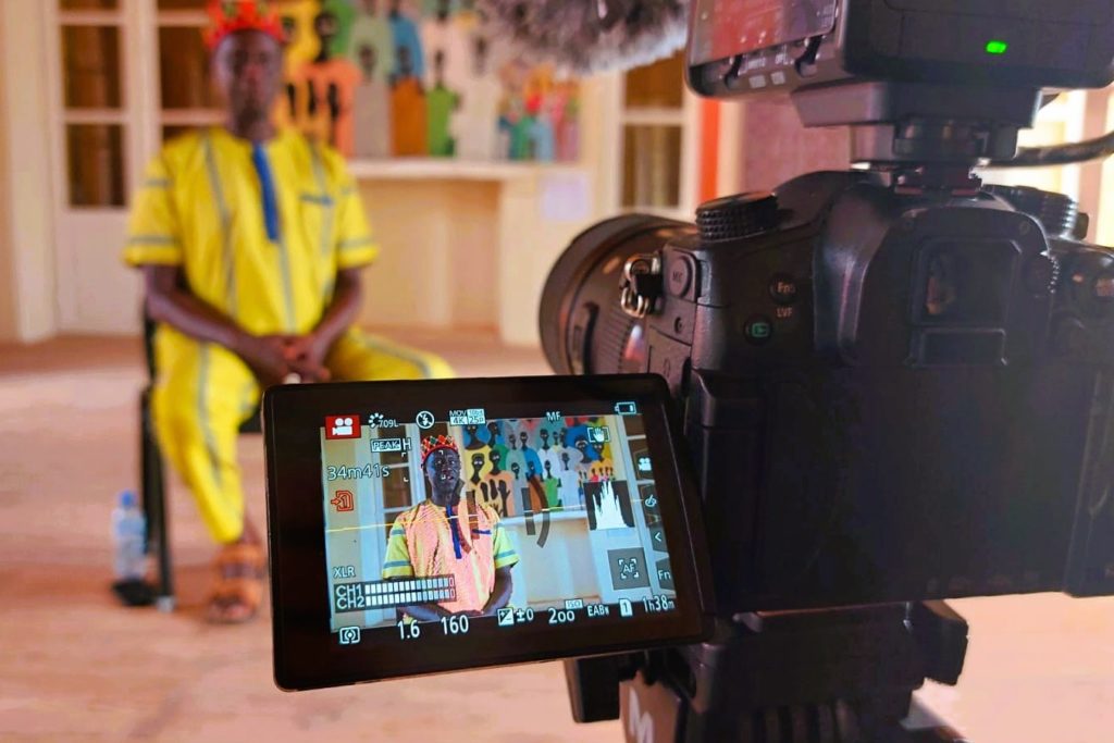 The video screen of a professional camera that is recording a man in a bright yellow outfit with a red and green cap on his head and a colorful painting of people behind him. 