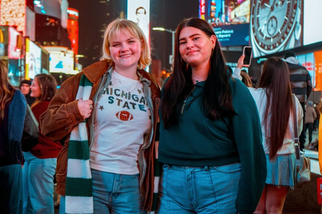 Two young women stand together in Times Square, smiling at the camera. One wears a brown jacket over a Michigan State t-shirt, and the other wears a dark green sweatshirt. They are surrounded by a crowd and bright digital billboards.