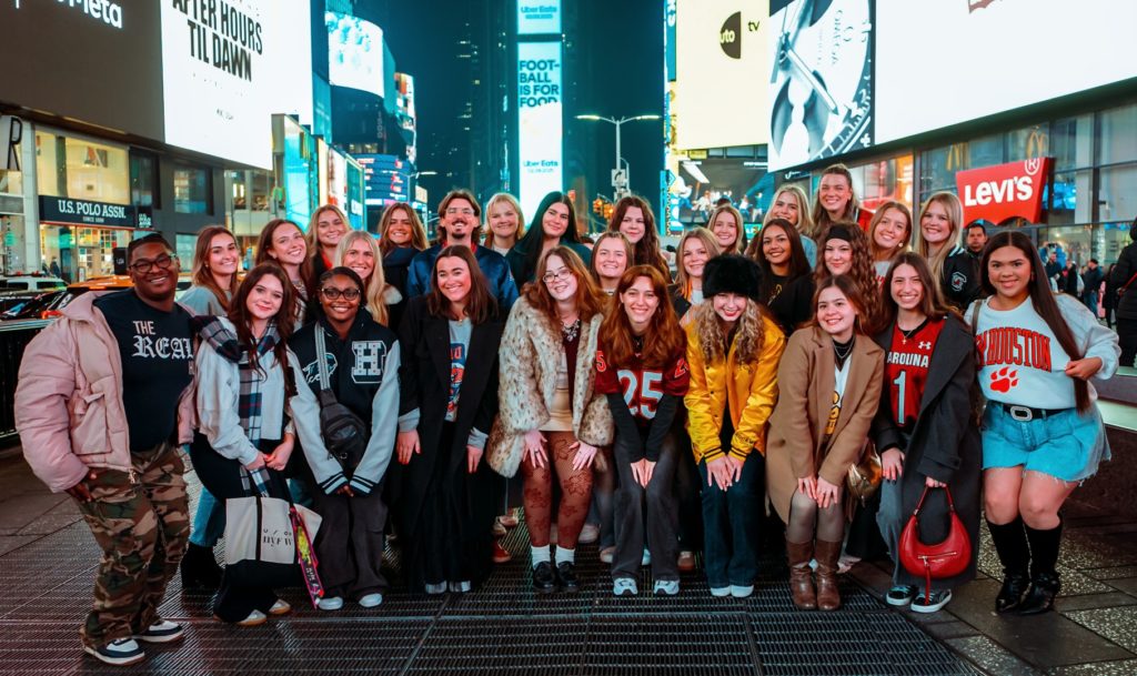 A large group of young people pose together in Times Square, dressed in a mix of casual and sporty attire. Neon billboards and city lights illuminate the background.