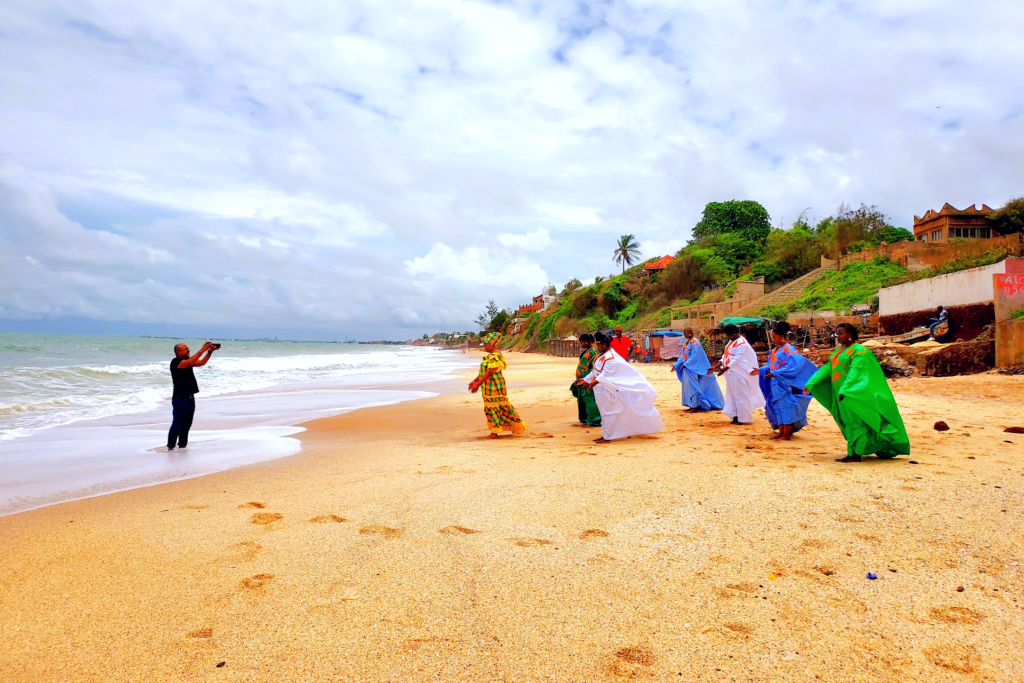 A man standing in the water at the edge of a beach holding a camera in the air pointed at a group of women in brightly colored dresses who are standing on the beach in a triangle formation.