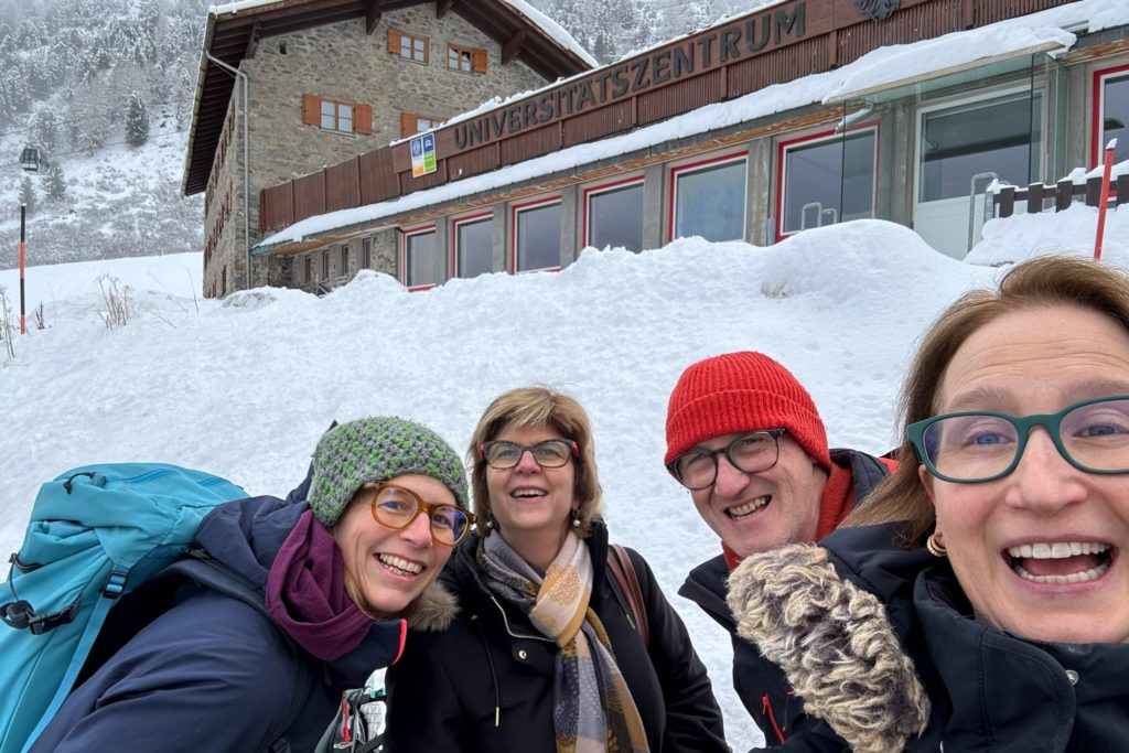 Four people (three women and one man) outside with a large snowbank behnd them and an alpine building with the word "Universitätszentrum" on it.