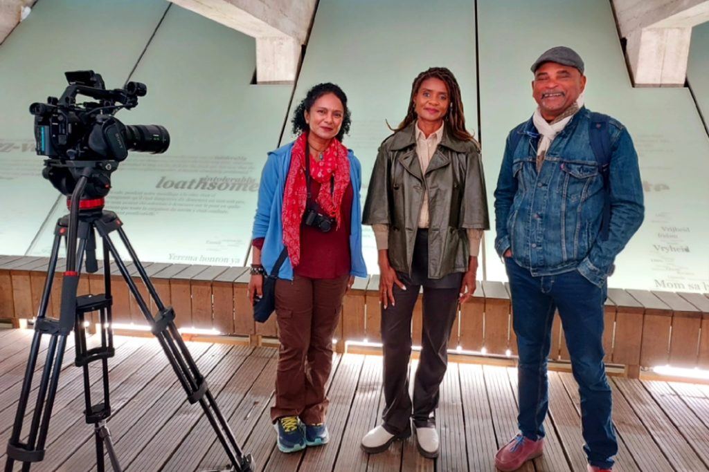 Three people stand in front of a slanted wall with text on it. A camera is set up on a tripod in the foreground. The person on the left wears a red scarf and carries a camera, the person in the middle wears a green jacket, and the person on the right wears a denim jacket and cap. They are all smiling.