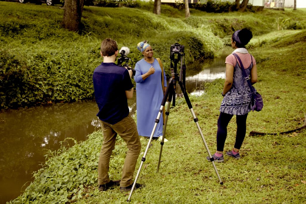 A woman wearing a blue dress and headwrap speaks while being filmed by a two-person camera crew in a lush, green outdoor setting. A small body of water runs alongside them. 