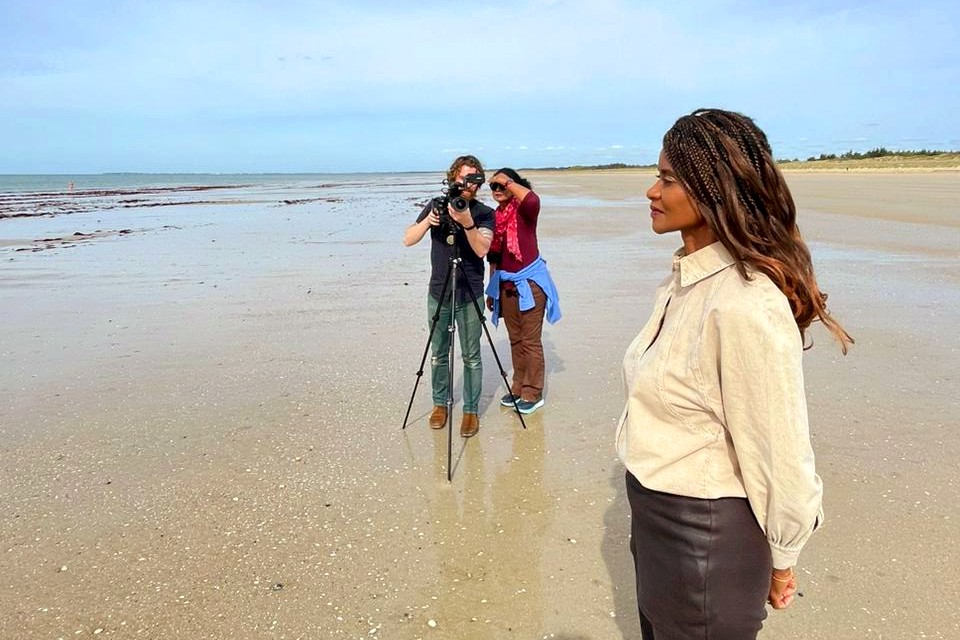  A woman with long braided hair stands on a sandy beach, gazing into the distance. Behind her, a person films with a tripod-mounted camera while another crew member watches. The ocean and horizon are visible in the background.