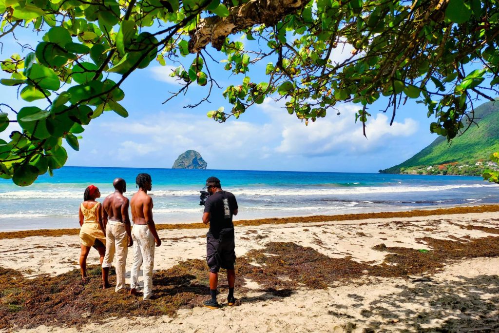 Three people (one woman and to men) standing on a very beautiful beach with another man video recording them. In the ocean there is a large rock and there is a very grrn mountain ide that alo can be seen that is rising from the ocean. 