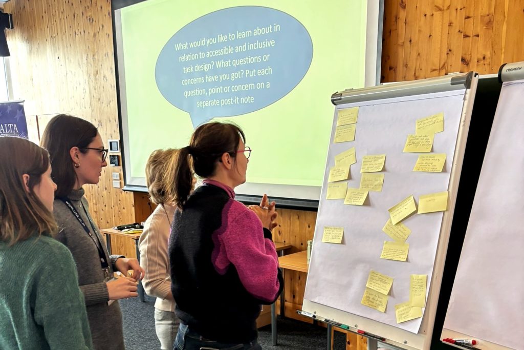 Four women looking at a large white piece of papers that has several post-it notes attached to it with writing on them. Behind then is a large screen with the following words on it: " What would you like to learn about in relation to accessible and inclusive task design? What questions of concerns have you got? Put each question, point or concern on a separate post-it note."