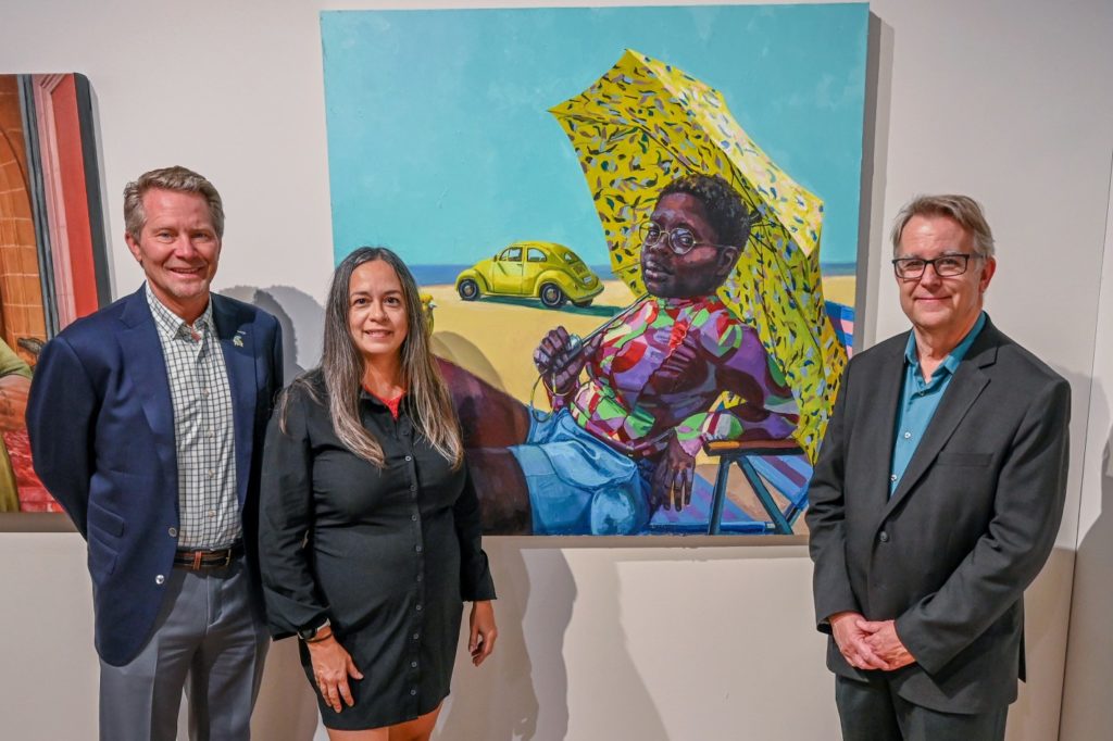 Three people - two men and one woman - standing in front of a colorful painting showing a Black woman sitting under a yellow umbrella on the beach with a yellow car in the background. 