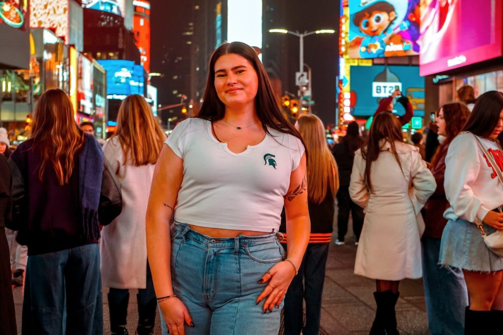 A young woman stands in Times Square, wearing a white cropped t-shirt with the Michigan State Spartans logo and blue jeans. She is surrounded by city lights and a bustling crowd.