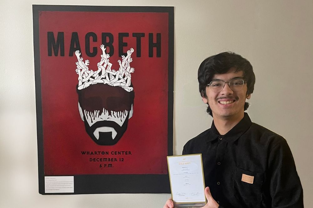 A picture of a man with a black shirt and glasses holding an award; he stands next to a red poster. 