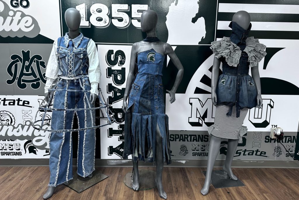 Three mannequins dressed in elaborate denim outfits stand in front of a wall featuring Michigan State University logos and the year "1855." The outfits incorporate various denim textures, patterns, and embellishments.