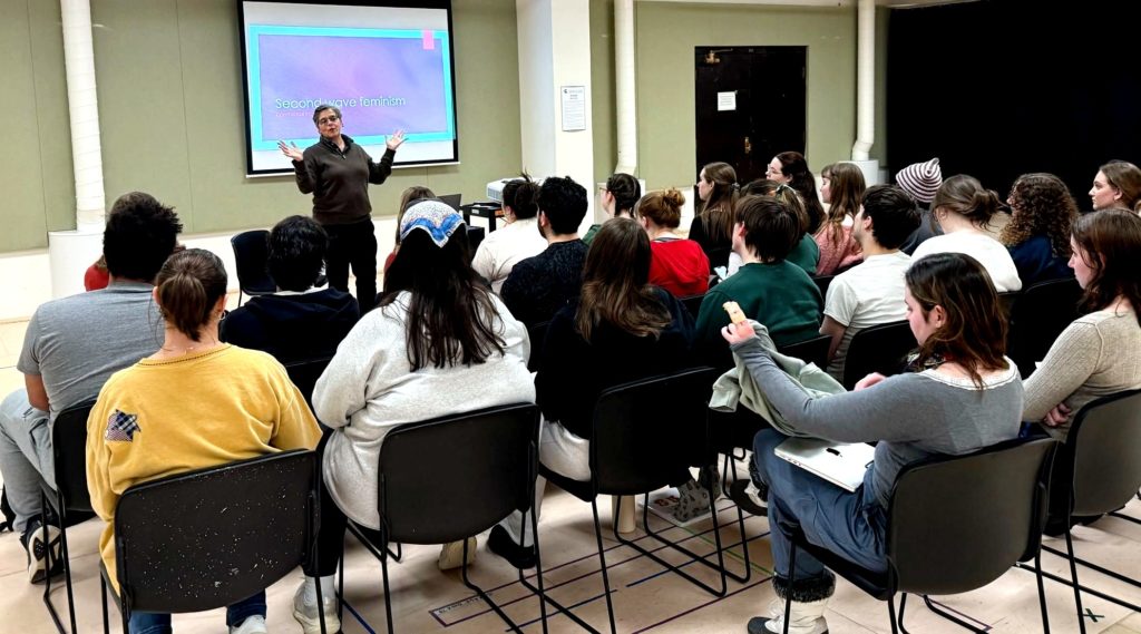 A woman with hands raised talking to a class full of students and behind her is a screen with the words "second-wave feminism" on it. 