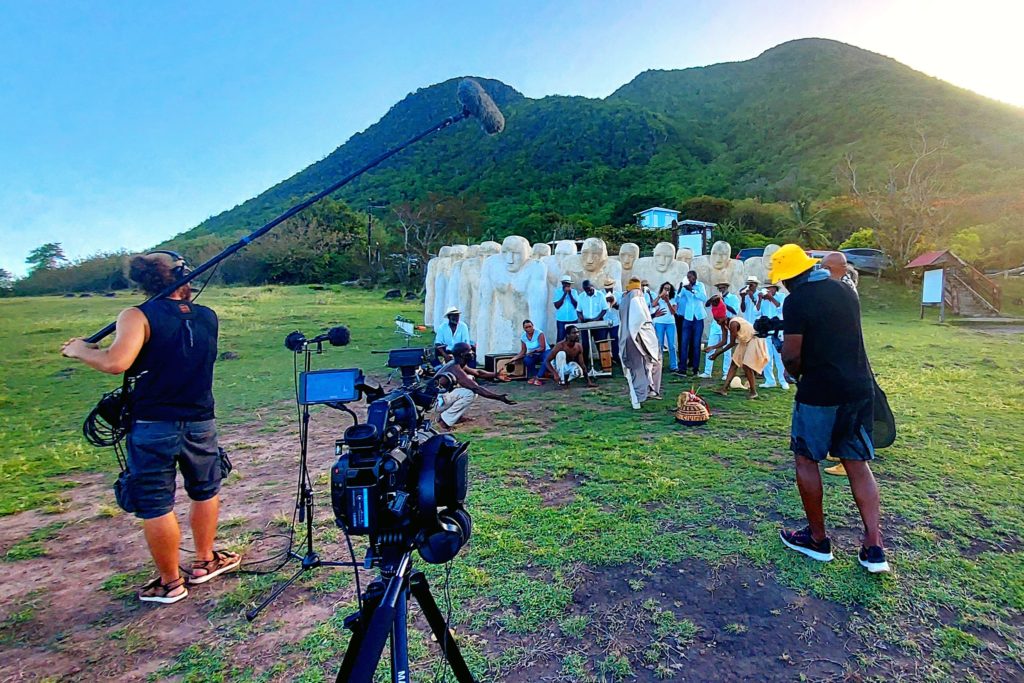 A film crew filming a group of people at the location of an outdoor sculpture, the Mémorial CAP 110.