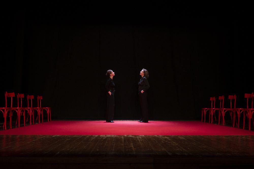 Two women on a theatre stage look at each other in the center. The stage is lit in red and empty chairs line the left and right sides of the stage. Photo by Federico Sigillo.