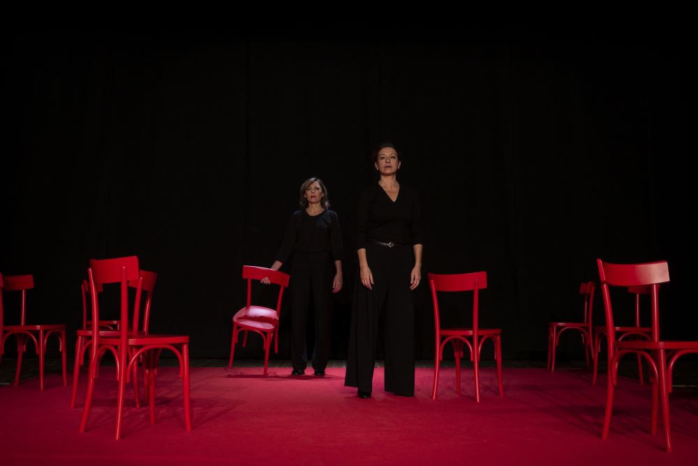 Two women stand in the center of a theatre stage looking straight ahead. One woman holds the back of a chair. The stage is lit in red and empty chairs surround the two women. Photo by Federico Sigillo.