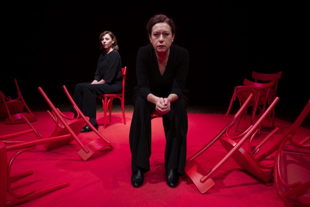 Two women wearing black sit on a theatre stage that is lit in red. One woman is sitting and looking straight ahead. The other woman is more in the background of the left and sitting at an angle. A group of discarded chairs surround both women. Photo by Federico Sigillo.