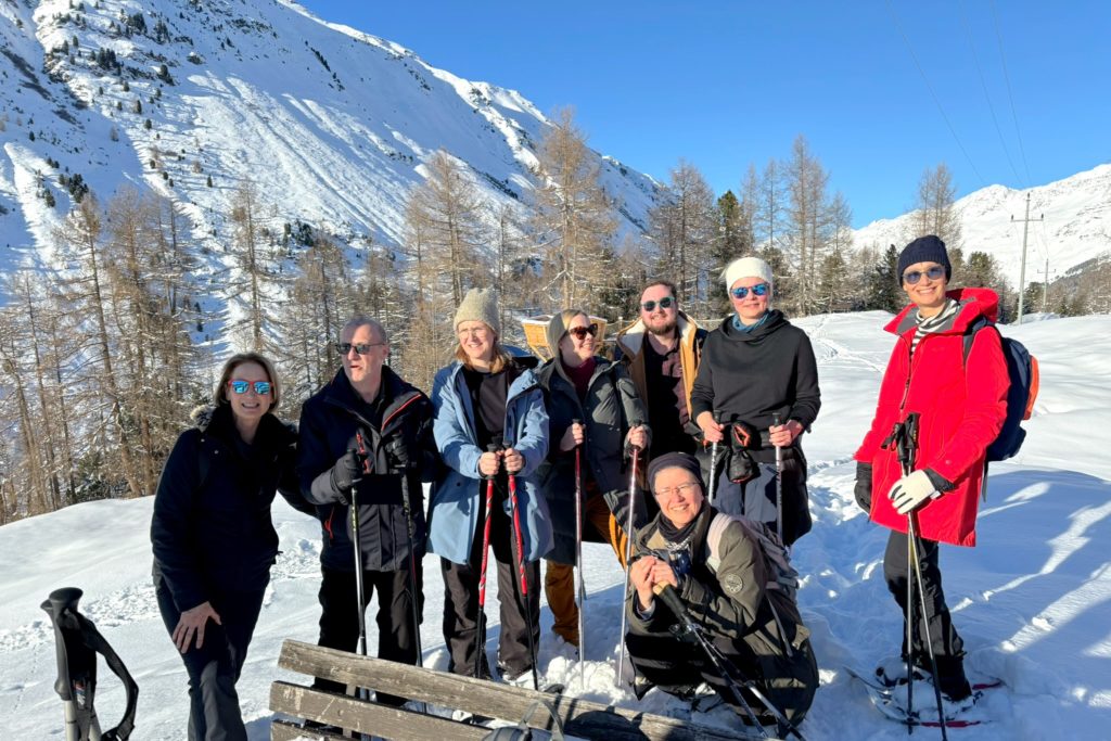 A group of eight people (six women and two men) all in winter coats and standing outside in the snow wearing snowshoes and holding onto poles for snowshoes. There are trees and a large hill in the background. 