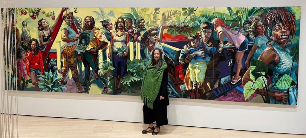 Woman standing in front of a large colorful painting, showing everal women of color doing various activities.