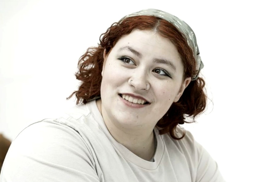 headshot of a woman with red, shoulder-length curly hair and wearing a head scar and two nose rings. 